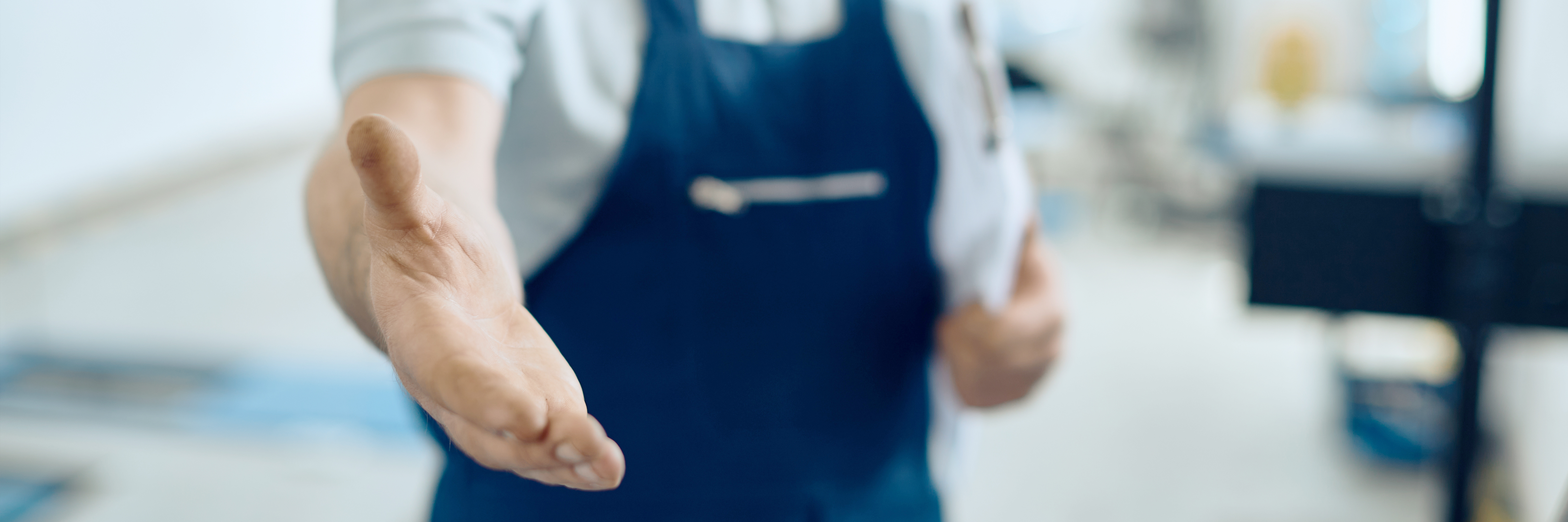 Close-up of a man's right hand extended to shake. The man is wearing blue dungarees and is carrying a white clipboard in the other hand.