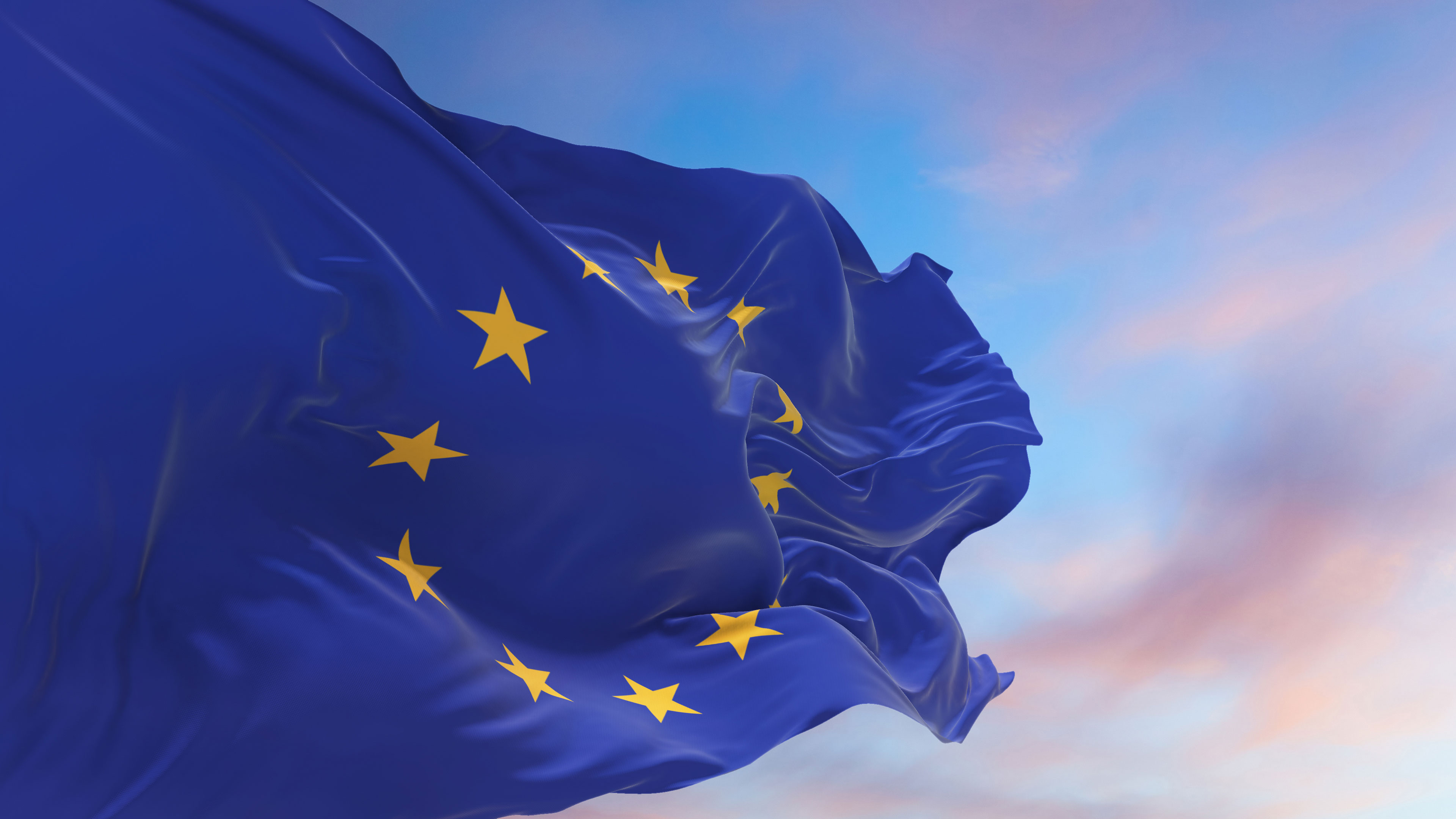 An EU flag flies in the wind on a flagpole during sunet. The sky in the background is slightly cloudy.